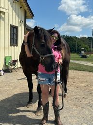 Smiling woman in a pink shirt, ripped denim shorts and cowboy boots hugging a large black horse by a yellow barn on a sunny summer day at a rural stable