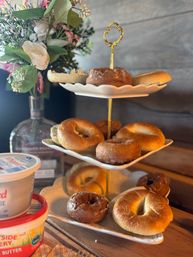 Three-tiered white-and-gold stand with assorted bagels and glazed donuts, tubs of cream cheese on a rustic wooden table and a floral vase — cozy brunch spread