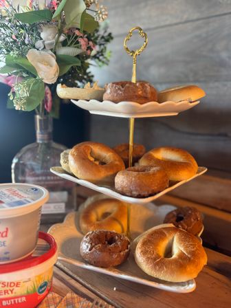 Three-tiered white-and-gold stand with assorted bagels and glazed donuts, tubs of cream cheese on a rustic wooden table and a floral vase — cozy brunch spread