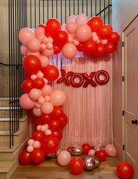 Valentine’s-themed indoor photo backdrop in a home entryway: pink fringe curtain framed by a red and pink balloon arch with scattered balloons and metallic red “XOXO” foil letters.