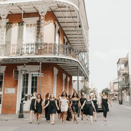 Group of women in black dresses, one wearing a white sash, walking hand-in-hand across a New Orleans French Quarter street under an ornate wrought-iron balcony