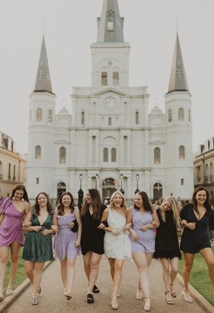 Eight women linked arm-in-arm, laughing and walking in colorful short dresses with a woman in a white dress, in front of St. Louis Cathedral at Jackson Square, New Orleans.