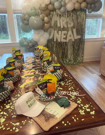 Bright indoor bridal shower setup with a long wooden table strewn with green confetti, gift baskets and a “Pickle Baller” hat, pastel sage and cream balloon garland and silver fringe backdrop
