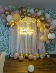 Indoor party backdrop with pastel pink, white and gold balloon garland, cream and blush streamers, neon sign reading 'TIL DEATH DO US PARTY', palm-leaf wallpaper and scattered balloons on a hardwood floor.