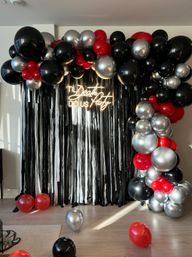 Indoor photo backdrop with black fringe curtain, large black, silver and red balloon arch and clusters, neon sign reading Til Death Do Us Party, and scattered balloons on a hardwood floor.