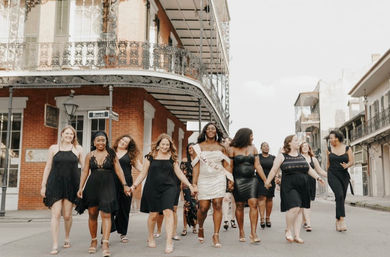Group of women in black dresses with a bride-to-be in white leading a joyful bachelorette walk down a New Orleans French Quarter street lined with brick buildings and ornate wrought-iron balconies.