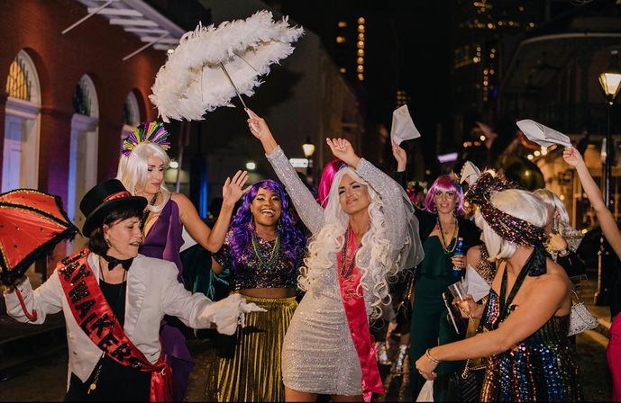 Joyful group in colorful wigs, sequins and beads dancing under a feathered parasol during a nighttime Mardi Gras–style street celebration