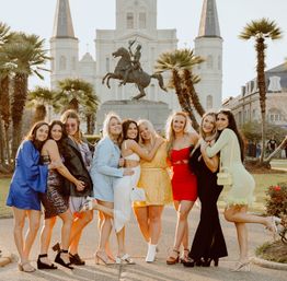 Nine friends in colorful party dresses and heels posing and laughing in front of the equestrian statue and St. Louis Cathedral at Jackson Square, New Orleans, with palm trees and warm sunset light.