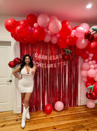 Woman in a white party dress and white cowboy boots posing indoors at a bachelorette party in front of a red metallic fringe backdrop with red and pink balloon arch and cherry foil balloons.