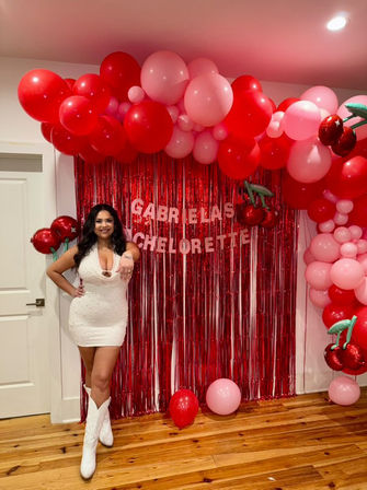 Woman in a white party dress and white cowboy boots posing indoors at a bachelorette party in front of a red metallic fringe backdrop with red and pink balloon arch and cherry foil balloons.