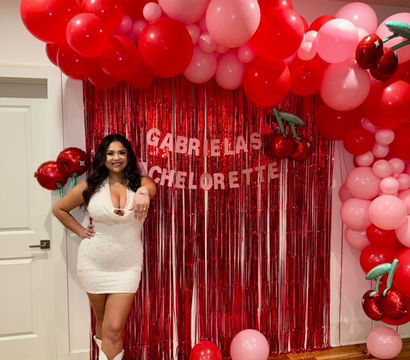 Woman in a white party dress and white cowboy boots posing indoors at a bachelorette party in front of a red metallic fringe backdrop with red and pink balloon arch and cherry foil balloons.