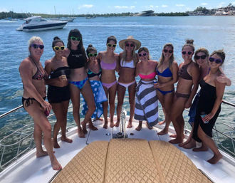 Group of women in bikinis and colorful sunglasses posing on the bow of a yacht at a sunny marina with boats and turquoise water, summer boat party vibe.