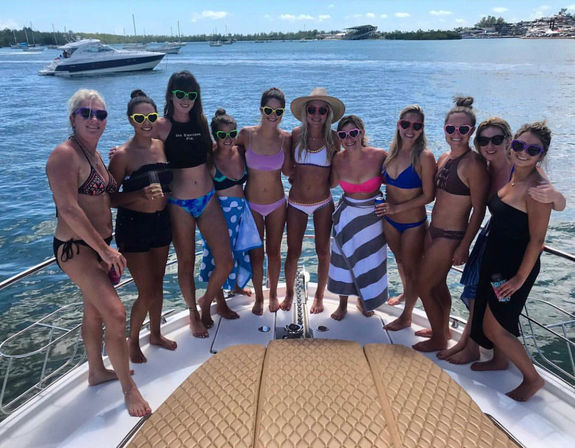 Group of women in bikinis and colorful sunglasses posing on the bow of a yacht at a sunny marina with boats and turquoise water, summer boat party vibe.