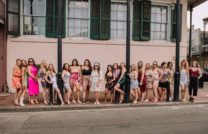 Large group of women in colorful party dresses posing on a sidewalk in front of a pastel building with green shutters and iron posts on a historic city street