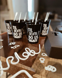 Black plastic cups reading 'TIL DEATH DO US PARTY' with white curly straws and confetti on a wooden table, spooky-chic wedding/bachelorette party decor.