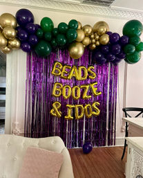 Indoor Mardi Gras–style party backdrop with purple metallic fringe curtain, green, purple and gold balloon garland and gold foil letter balloons, cream sofa with pink pillow and wooden floor in foreground.