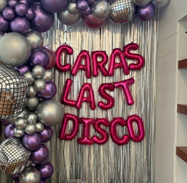Disco-themed indoor party backdrop with silver fringe curtain, metallic purple and silver balloon garland, mirrored disco-ball decorations, and hot-pink foil letter balloons forming a personalized “Last Disco” message.