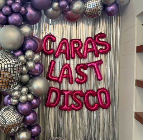 Disco-themed indoor party backdrop with silver fringe curtain, metallic purple and silver balloon garland, mirrored disco-ball decorations, and hot-pink foil letter balloons forming a personalized “Last Disco” message.