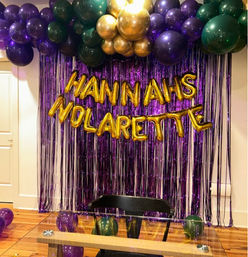 Mardi Gras–style purple, green and gold balloon arch over a purple metallic fringe backdrop with gold letter balloons spelling a celebratory message, glass table and chair in the foreground.