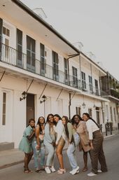 Seven women smiling and posing on a sunny historic city street in front of a white building with wrought-iron balconies — casual urban lifestyle group photo.