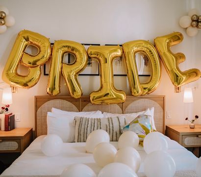 Bridal suite bedroom decorated with large gold foil balloons spelling BRIDE above a bed scattered with white balloons and neutral pillows