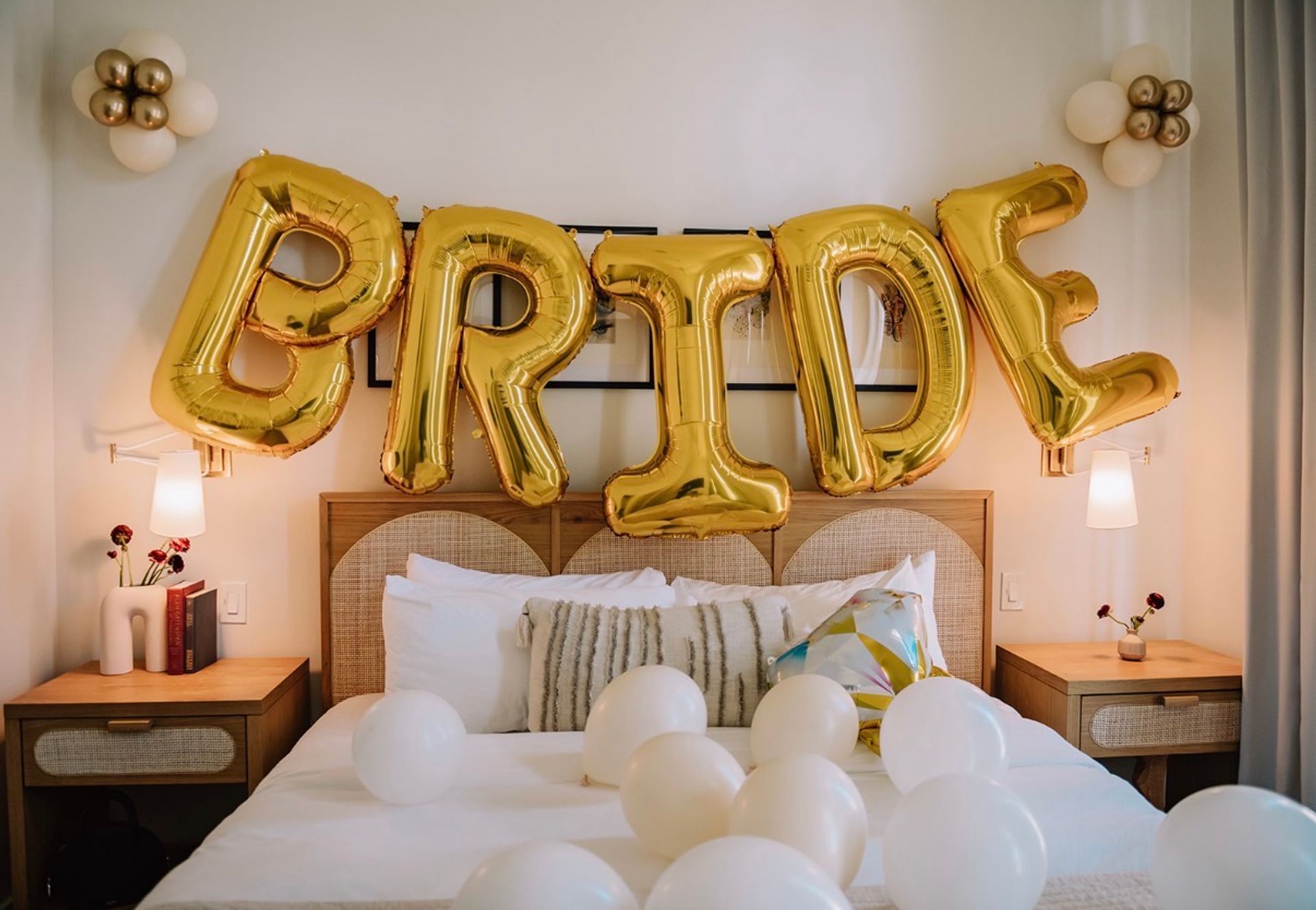 Bridal suite bedroom decorated with large gold foil balloons spelling BRIDE above a bed scattered with white balloons and neutral pillows