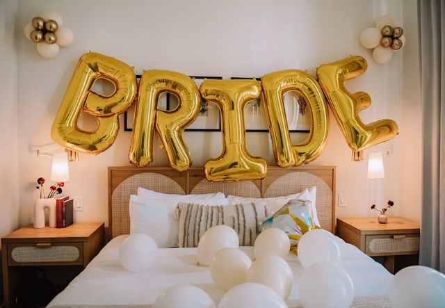 Bridal suite bedroom decorated with large gold foil balloons spelling BRIDE above a bed scattered with white balloons and neutral pillows