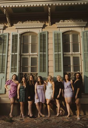 Eight women in colorful dresses posing on a brick sidewalk in front of a sunlit pastel Creole-style building with mint shutters and arched windows