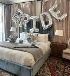 Bridal bedroom scene with silver foil "BRIDE" balloons over a velvet headboard, neutral linens, mirrored disco-ball cube decor on the bed, bedside lamps and patterned area rug
