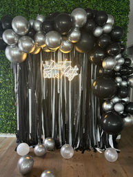 Indoor photo booth backdrop with black and silver balloon arch, black-and-white streamers, glowing neon sign on a green hedge wall and scattered balloons on a wood floor