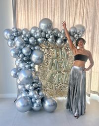 Smiling woman posing with arm raised beside a large metallic silver balloon arch and gold sequin backdrop featuring letter balloons reading "IN HER ERA", wearing a black bandeau top and silver pleated skirt at a celebratory event.