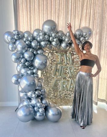 Smiling woman posing with arm raised beside a large metallic silver balloon arch and gold sequin backdrop featuring letter balloons reading "IN HER ERA", wearing a black bandeau top and silver pleated skirt at a celebratory event.