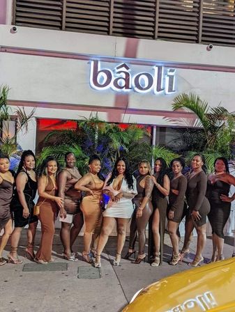 Group of women in evening dresses posing for a night-out photo outside a lit-up, tropical-themed restaurant entrance with neon signage and palm plants