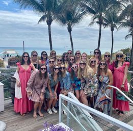 Large group of women in colorful sundresses and sunglasses posing on a beachside wooden deck with palm trees, ocean waves, and a partly-cloudy sky in the background.