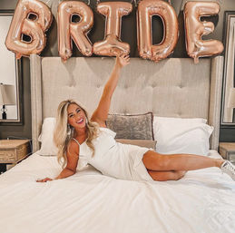 Smiling woman in a white dress lounging on a bed beneath rose-gold 'BRIDE' balloons for a bridal celebration in a cozy bedroom