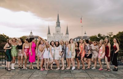 Large group of women in colorful party dresses laughing and posing on a plaza with St. Louis Cathedral and an American flag in Jackson Square, New Orleans, at sunset