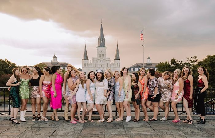 Large group of women in colorful party dresses laughing and posing on a plaza with St. Louis Cathedral and an American flag in Jackson Square, New Orleans, at sunset