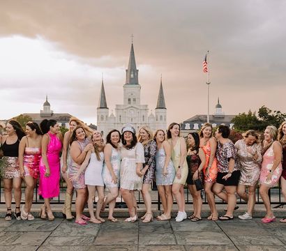 Large group of women in colorful party dresses laughing and posing on a plaza with St. Louis Cathedral and an American flag in Jackson Square, New Orleans, at sunset