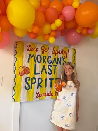 Smiling woman holding an orange spritz cocktail in front of a hand-painted yellow-and-white party sign beneath a bright pink, orange and yellow balloon garland.