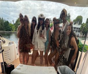 Group of friends posing for a selfie behind a wet glass panel on a yacht, wearing swimwear, cover-ups and sunglasses with a tree-lined waterfront and dockside homes in the background.