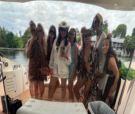 Group of friends posing for a selfie behind a wet glass panel on a yacht, wearing swimwear, cover-ups and sunglasses with a tree-lined waterfront and dockside homes in the background.