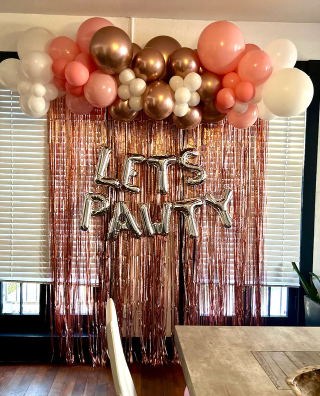 Indoor party backdrop with rose-gold metallic fringe curtain, silver letter balloons spelling "LET'S PARTY" and a pink, rose-gold and white balloon garland above a dining table.