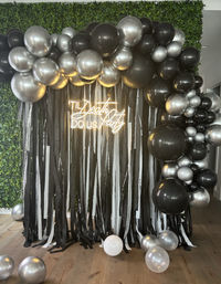 Black-and-silver balloon arch with metallic balloons and black-and-white streamer backdrop, neon sign reading 'Til Death Do Us Party' on a green hedge wall — chic wedding/party photo booth decor.