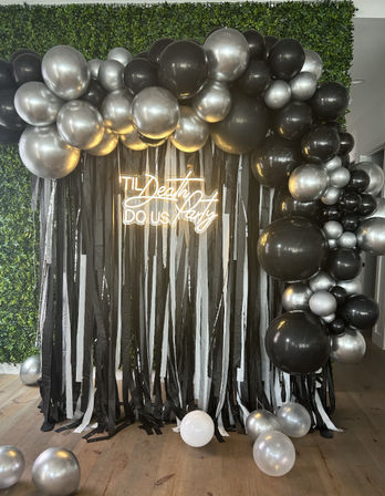 Indoor wedding photo-backdrop with black and metallic silver balloon garland, black-and-white streamers and neon sign reading 'Til Death Do Us Party' against a faux boxwood wall.