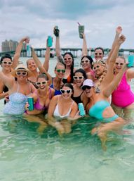 Cheerful group of friends in colorful swimsuits and sunglasses posing and raising canned drinks in shallow turquoise beach water with a bridge and buildings in the background — lively summer seaside celebration.