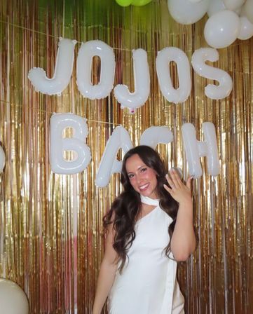 Smiling woman in a white dress flashing an engagement ring at a bridal shower photo booth with white letter balloons and a gold fringe backdrop.