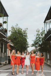 Seven women — six in red dresses and one in a white dress — link arms and smile on a crosswalk in a historic New Orleans French Quarter street framed by wrought-iron balconies and hanging plants.