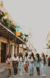 Seven friends laughing and walking arm‑in‑arm down a sunny New Orleans French Quarter street, passing ironwork balconies hung with flowers and an Irish shamrock flag.