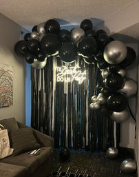 Indoor living-room party backdrop with a dramatic black-and-silver balloon arch over metallic fringe curtains, glowing white neon script sign, gray couch to the side and a few balloons on the carpet — moody monochrome celebration decor.