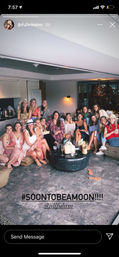 Large group of women in a bright modern lounge, wearing summer dresses and hats, smiling and holding drinks around a low coffee table with straw bags — fun bachelorette-style celebration indoors.